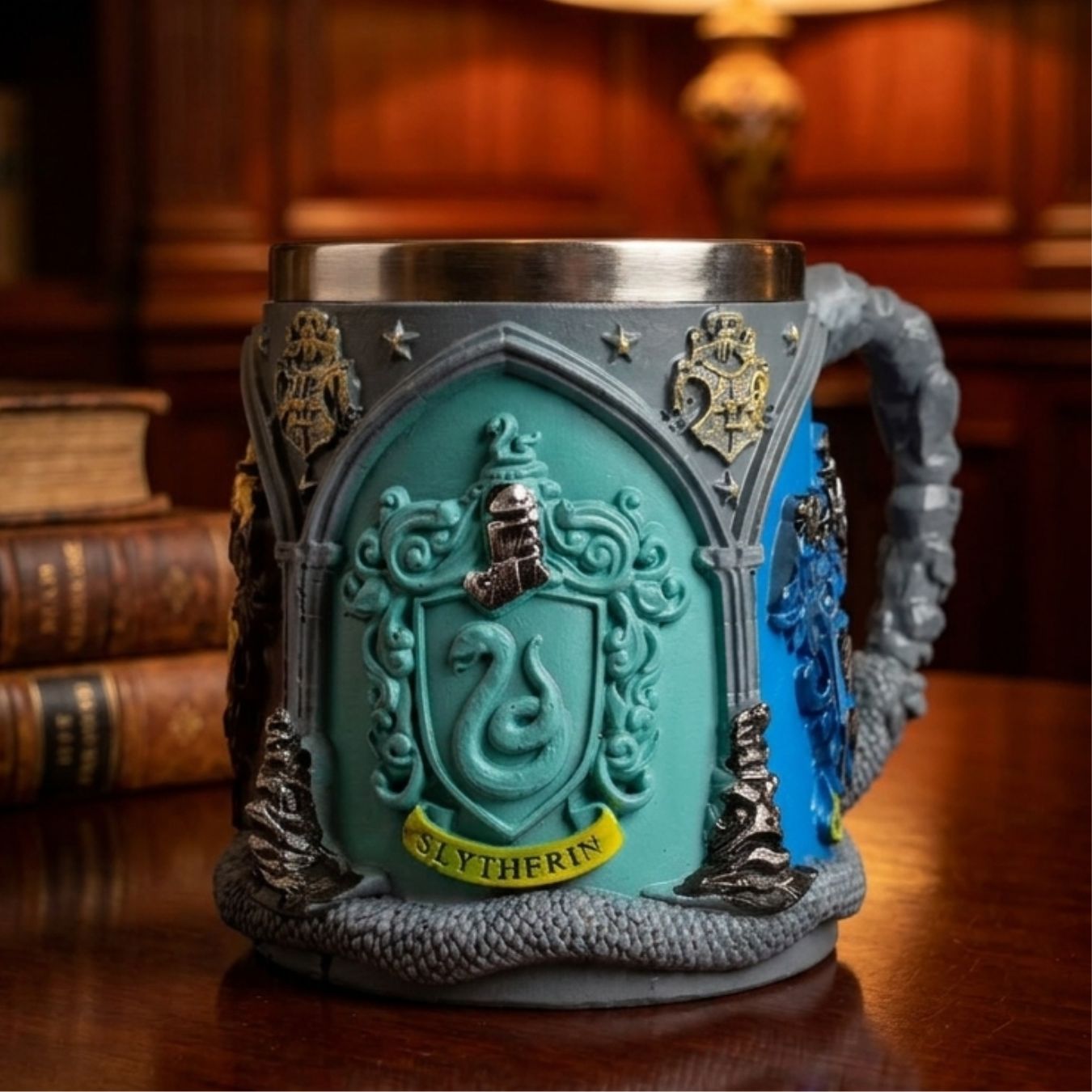 A Slytherin themed mug with a textured gray stone-like body, a turquoise center panel featuring a snake crest and the word 'SLYOTHERIN', blue side detailing, and a silver metal rim, shown on a wooden table with books and a lamp in the background.
